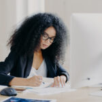 Photo of serious Afro woman writes in papers, sits at table with modern computer, creats article in newspaper, wears glasses and eyewear, poses indoor, works remotely. People and job concept